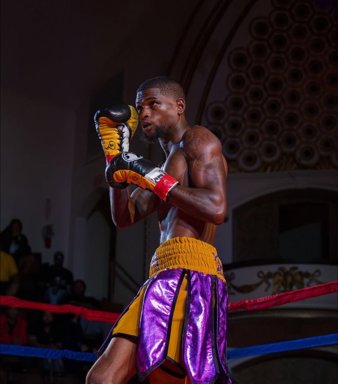 A boxer in purple and gold trunks holding his guard under the ring lights